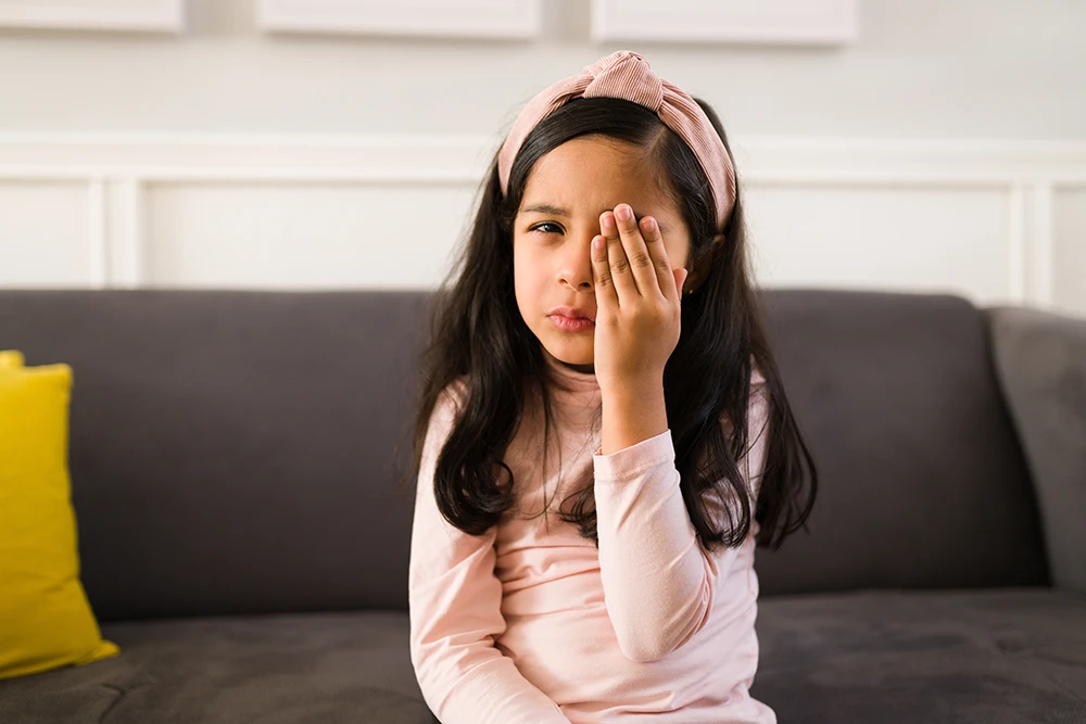 a little girl sitting on a couch and holding a hand over her eye