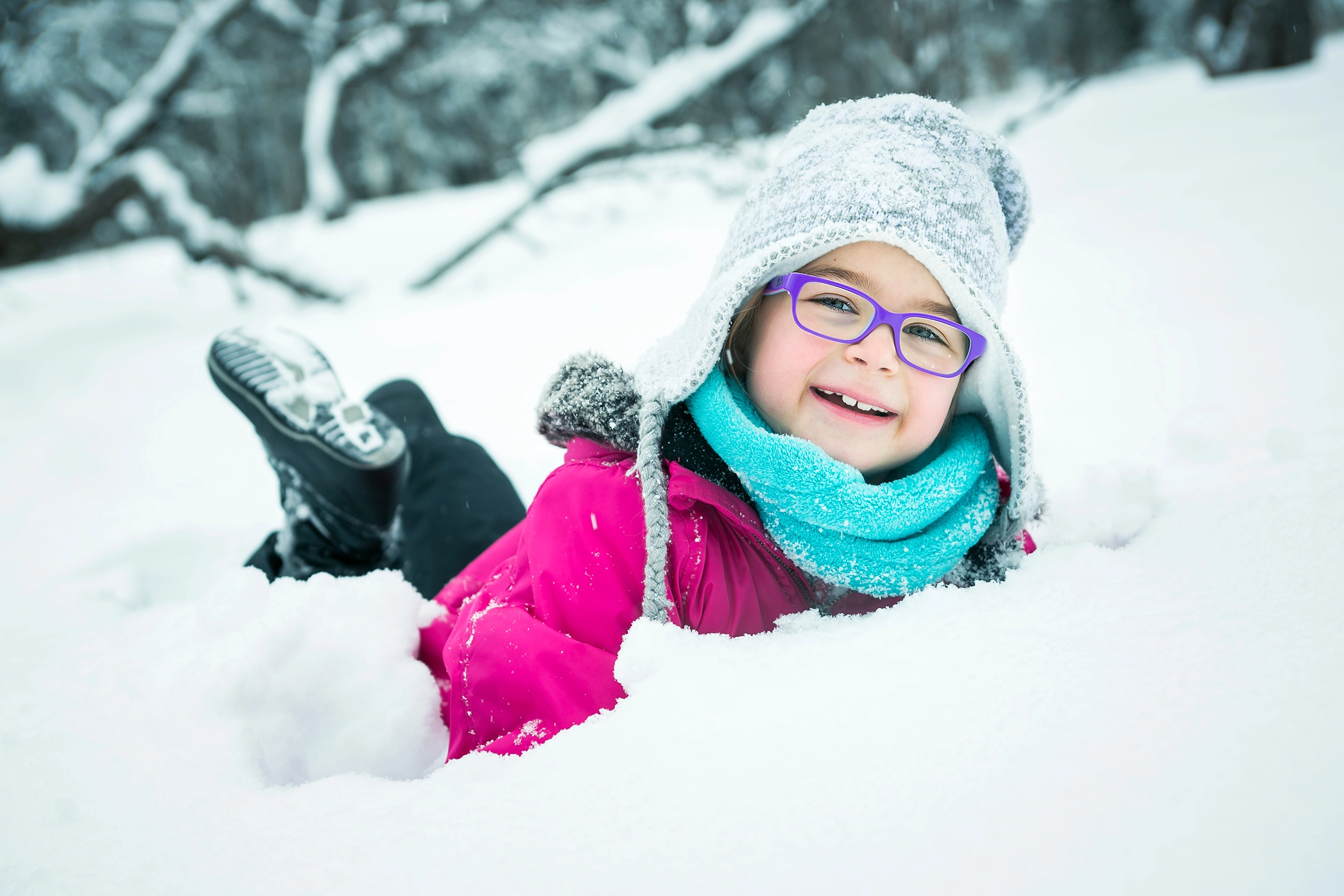 A little girl in winter clothes and purple framed glasses plays in the snow.